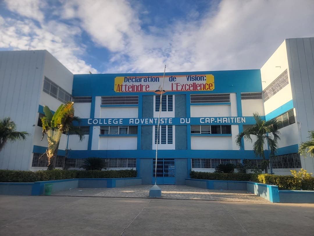 Façade bleu ciel et blanche du Collège Adventiste du Cap-Haïtien sous un ciel nuageux, avec la bannière « Déclaration de Vision : Atteindre l'Excellence », la cour et les palmiers devant l'entrée.